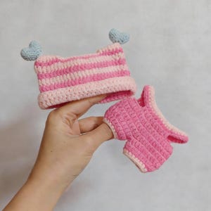 May include: A crocheted set consisting of a pink and white striped hat with two light blue heart-shaped accents on top and a pair of matching pink crocheted shorts with a light pink trim along the edges of the waistband and leg openings.
