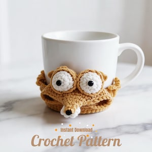 May include: A white ceramic mug with a crocheted fox-like cozy. The cozy is tan with white and black accents, featuring large eyes and a snout. The text "Instant Download Crochet Pattern" is at the bottom.