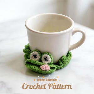May include: A white ceramic mug rests on a green crocheted frog-shaped coaster. The coaster features white eyes with black crosses, a pink mouth, and the text "Instant Download Crochet Pattern" in orange and brown.