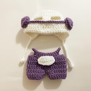 May include: A handmade crochet baby outfit in white and purple. The set includes a helmet-style hat with ear flaps and a pair of shorts. The hat has purple accents and the shorts feature a white accent. The outfit is designed for a baby.
