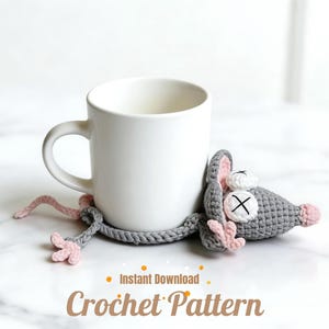 May include: A white ceramic mug rests next to a gray crocheted mouse coaster with pink accents. The mouse has a white X on its eye and pink ears and feet. The words "Instant Download Crochet Pattern" are below.