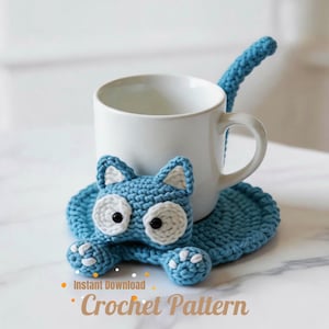 Puede incluir: Un posavasos de gato de ganchillo azul con detalles blancos, sosteniendo una taza blanca. El posavasos tiene una cara de gato, patas y una cola. El texto "Crochet Pattern" es visible.