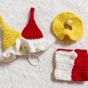 May include: Crocheted baby accessories in yellow, red, and white. The set includes two hats, a circular bib, and a pair of booties. The items are handmade and placed on a white, textured surface.