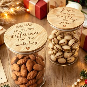 Op de afbeelding: Twee glazen voorraadpotten met ronde bamboe deksels. Het linker deksel is gegraveerd met "never FORGET THE difference THAT YOU make lucy" en een hartje. Het rechter deksel is gegraveerd met "papa WE TRY OUR BEST mostly LOVE jane" en een hartje.