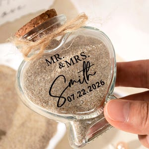 Peut inclure: Une bouteille en verre en forme de cœur remplie de sable beige, dotée d'un bouchon en liège attaché avec de la ficelle. Le verre porte l'inscription noire MR. & MRS. Smith 07.22.2026. La bouteille est transparente et a une silhouette en cœur.