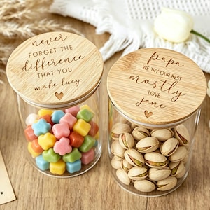 May include: Two clear glass jars with round bamboo lids. The left lid is engraved with "never FORGET THE difference THAT YOU make lucy" and a heart. The right lid is engraved with "papa WE TRY OUR BEST mostly LOVE jane" and a heart. The jars contain colorful candies and pistachios.