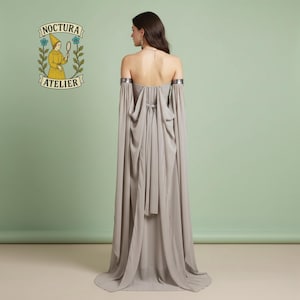 May include: A floor-length, strapless, light gray gown featuring long, flowing detached sleeves with metallic silver arm cuffs. The back of the dress includes a tie closure and draped fabric panels. The Noctura Atelier logo is visible in the corner.