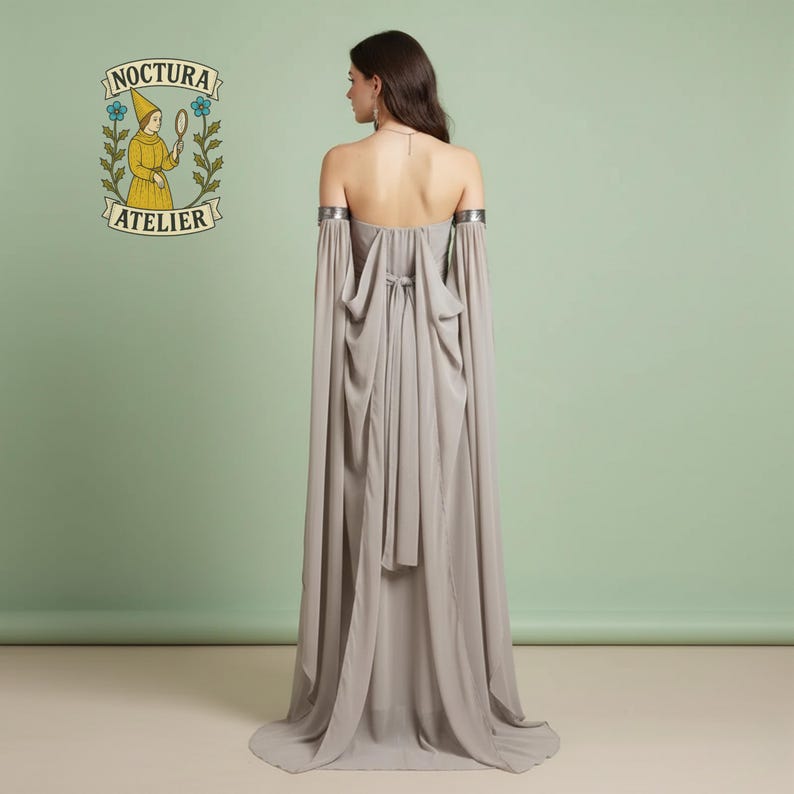 May include: A floor-length, strapless, light gray gown featuring long, flowing detached sleeves with metallic silver arm cuffs. The back of the dress includes a tie closure and draped fabric panels. The Noctura Atelier logo is visible in the corner.