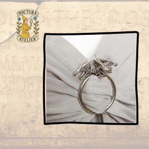 May include: A silver-toned metal scarf ring featuring two detailed dragon heads facing outward, securing a piece of light gray, finely pleated fabric. The accessory has a circular band design that holds the fabric in place through the center.