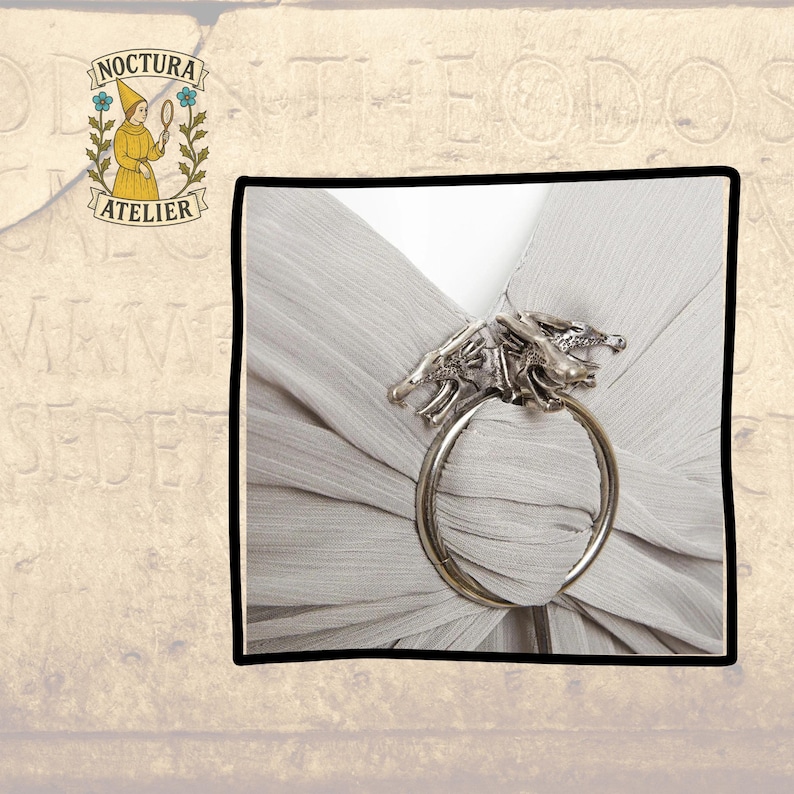 May include: A silver-toned metal scarf ring featuring two detailed dragon heads facing outward, securing a piece of light gray, finely pleated fabric. The accessory has a circular band design that holds the fabric in place through the center.