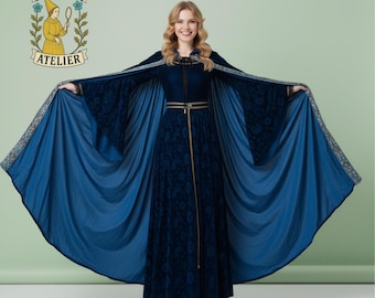 May include: A long, dark blue velvet dress with a matching cape. The cape has a lighter blue lining and decorative trim. The dress features a gold-colored belt and a high neckline. The image includes the text "NOCTURA ATELIER" with a logo.