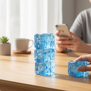May include: Three blue, square-shaped ice cubes with white floral patterns are stacked on a wooden table. A fourth ice cube is held by a person's hand. A person is using a smartphone in the background.
