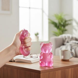 Puede incluir: Una bola antiestrés con forma de oso de goma rosa se sostiene en una mano, junto a una figura de oso de goma rosa. La bola antiestrés está siendo apretada, y la figura está sobre una mesa de madera. Una taza de té, gafas y un libro abierto también están sobre la mesa.