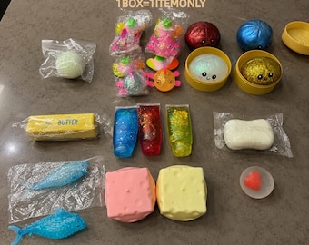 Pode incluir: Uma coleção de vários brinquedos sensoriais, incluindo pequenos peixes de plástico, recipientes de slime com glitter, cubos macios e recipientes redondos com rostos sorridentes. O texto SURPRISE BOX e 1BOX=1ITEMONLY é visível.