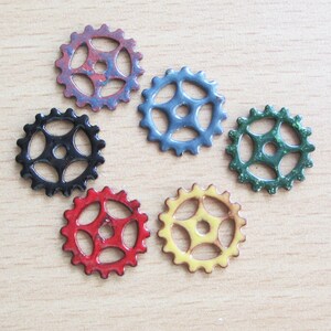 May include: A collection of colorful metal gear-shaped embellishments. The gears are in various colors, including red, black, blue, green, and yellow. Each gear has a central hole and multiple outer teeth. They are arranged on a light wood surface.