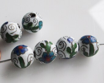Könnte beinhalten: Runde, weiße Perlen mit blauen, grünen und roten Blumenmustern. Die Perlen sind auf einem dünnen Metalldraht aufgefädelt. Die floralen Designs umfassen Wirbel und Blätter.