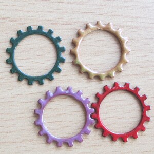 May include: Four colorful gear-shaped embellishments in green, tan, purple, and red. Each gear has a circular design with evenly spaced teeth around the outer edge. The gears are arranged on a light brown wooden surface.