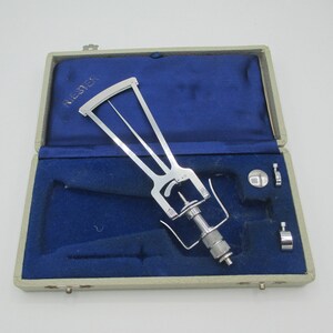 Könnte beinhalten: Ein silberfarbenes zahnmedizinisches Instrumentenset in einem blau samtgefütterten Etui. Das Set enthält ein Hauptinstrument mit gebogener Oberseite und verstellbaren Komponenten sowie mehrere kleinere Teile. Das Etui ist hellgrau mit dem Aufdruck "KIE-SYEK".