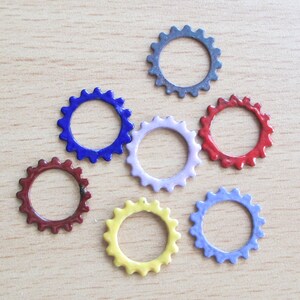 May include: A collection of colorful, small, gear-shaped embellishments in various colors, including blue, red, yellow, and purple. These decorative elements are arranged on a light wood-grain surface.