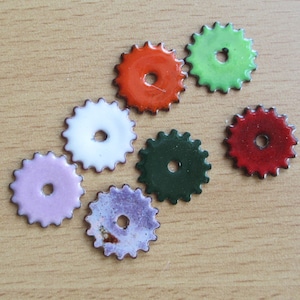 May include: A collection of colorful, enamel-coated metal gear-shaped components. The gears are in various colors, including red, green, white, pink, purple, and dark green. Each gear has a central hole and a scalloped edge.