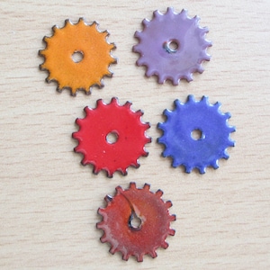 May include: Five colorful, enamel-coated gear-shaped components. The gears are in shades of orange, purple, red, blue, and a reddish-brown. Each gear has a central hole and a dark outline, set against a light wood grain background.