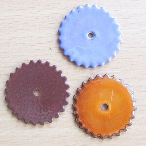 May include: Three colorful, round, ceramic pieces with a scalloped edge and a central hole. One is a muted blue, another is a deep brown, and the third is a vibrant orange. These decorative elements could be used for various craft projects.