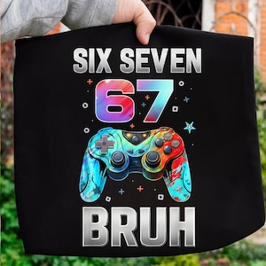 Puede incluir: Camiseta negra con la frase "SIX SEVEN 67 BRUH" en blanco. El número 67 tiene un degradado de azul, morado y rojo. Debajo hay un mando de videojuegos colorido.