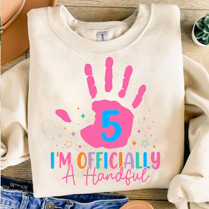 Op de afbeelding: Crèmekleurige sweatshirt met een grote roze handafdruk en het nummer 5 in blauw. De tekst "I'M OFFICIALLY A Handful" staat eronder in verschillende kleuren. Het ontwerp bevat kleurrijke steraccenten.