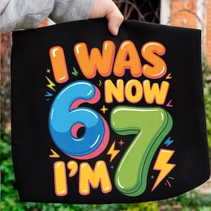 Puede incluir: Bolso tote negro con el texto "I WAS NOW 67 I'M" en naranja, azul y verde. Los números 6 y 7 son grandes y coloridos, con acentos de rayos. El bolso lo sostiene una persona.