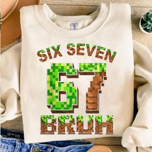 K&ouml;nnte beinhalten: Cremefarbenes Sweatshirt mit dem Text "SIX SEVEN BRUH" in einer pixeligen Schriftart. Die Zahlen "67" sind in einem gr&uuml;nen und braunen Pixeldesign gehalten. Ein l&auml;ssiges, bequemes Oberteil f&uuml;r den Alltag.