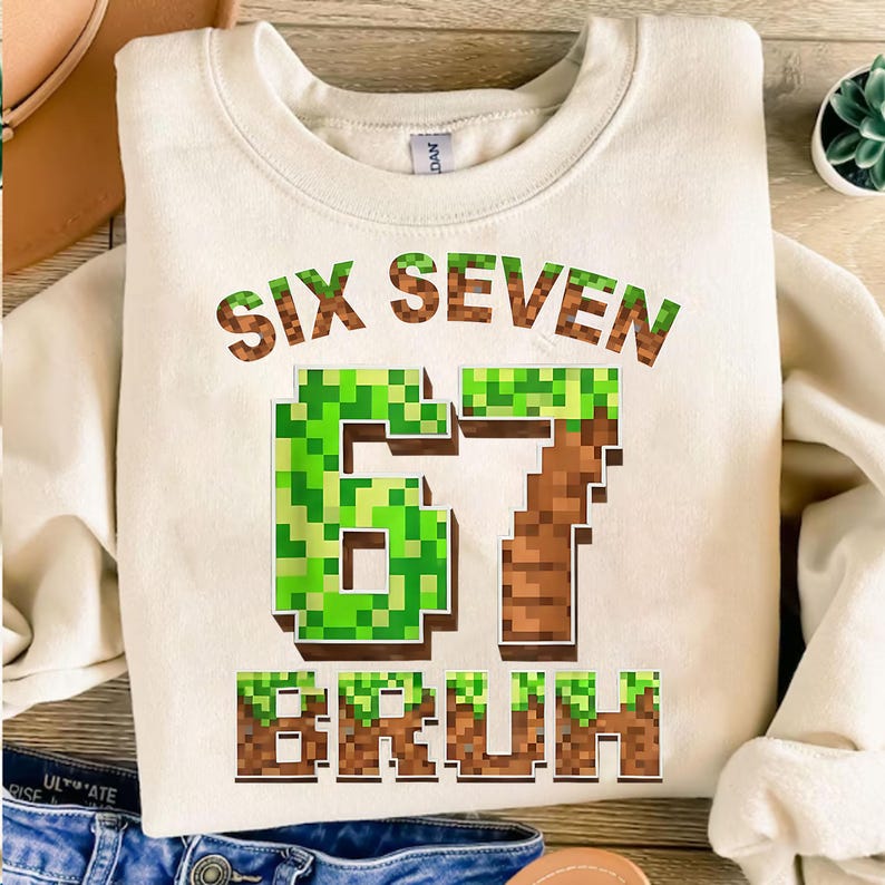 K&ouml;nnte beinhalten: Cremefarbenes Sweatshirt mit dem Text "SIX SEVEN BRUH" in einer pixeligen Schriftart. Die Zahlen "67" sind in einem gr&uuml;nen und braunen Pixeldesign gehalten. Ein l&auml;ssiges, bequemes Oberteil f&uuml;r den Alltag.