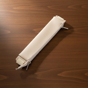 May include: A cream-colored, rectangular pencil case with a zipper closure and a small elastic strap. The case is made of a smooth material and is lying on a dark wood surface.