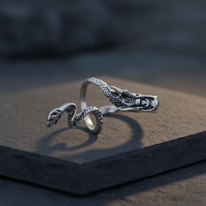 May include: A silver-colored ring in the shape of a snake. The snake's head is detailed with scales and a slightly open mouth. The snake's body wraps around the finger, with the tail end near the head. The ring is set against a dark background.