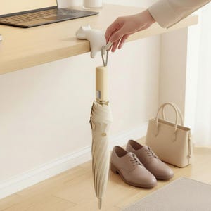 May include: A beige umbrella hanging from a white cat-shaped hook on a wooden desk. A pair of brown shoes, a beige handbag, and a laptop are also visible. The umbrella is closed and has a spiral design.
