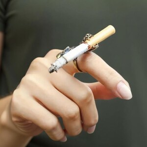 May include: A gold-toned cigarette ring holds a lit cigarette. The ring has an ornate design with a small blue accent. The cigarette is lit and has a gray ash. The background is a solid, muted green.