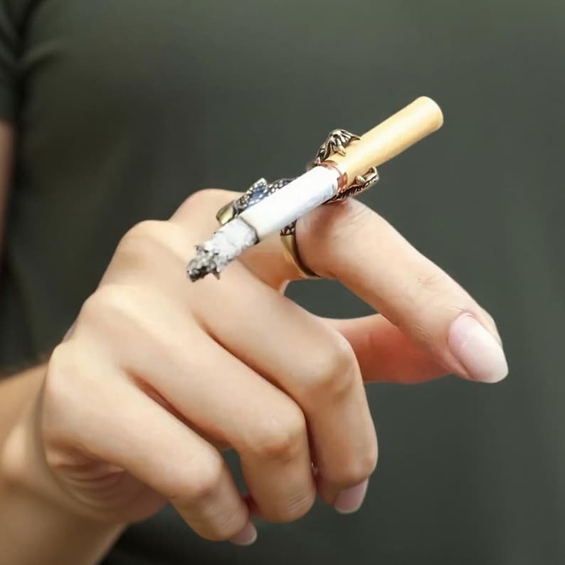 May include: A gold-toned cigarette ring holds a lit cigarette. The ring has an ornate design with a small blue accent. The cigarette is lit and has a gray ash. The background is a solid, muted green.