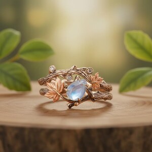 Puede incluir: Un conjunto de anillos de oro rosa con un diseño de ramas y acentos de hojas de arce. El anillo tiene una piedra preciosa azul claro en forma de pera y pequeñas piedras preciosas transparentes. El anillo está sobre una superficie de madera con un fondo verde borroso.