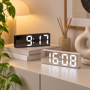 May include: Two rectangular digital clocks with mirrored surfaces and bright white LED displays. One clock shows the time 9:17, while the other displays 16:08. The devices feature a sleek, minimalist design with clear, large numerical digits.