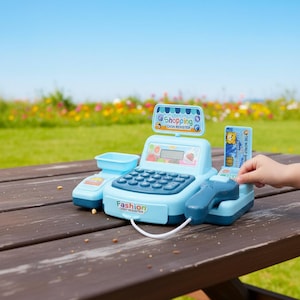 May include: A light blue toy cash register with a keypad, display, and scanner. The register has the word "Shopping" and "Fashion" printed on it. A child's hand is inserting a credit card into the register.