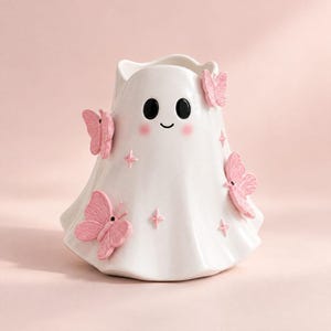 May include: A white ceramic ghost-shaped vase featuring a smooth finish, two black oval eyes, a small curved mouth, and soft pink blush marks. The vase is decorated with three-dimensional light pink butterflies and small pink star-shaped accents.