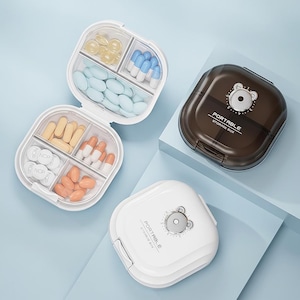 Puede incluir: Dos pastilleros portátiles, uno blanco y otro marrón. El pastillero blanco está abierto, revelando compartimentos llenos de pastillas. El pastillero marrón está cerrado y tiene un diseño de oso con el texto "PORTABLE STORAGE BOX".