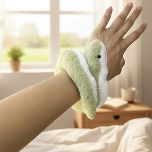 Könnte beinhalten: Ein grünes und weißes schlangenförmiges Armband aus weichem Plüschmaterial. Das Spielzeug ist um ein Handgelenk gewickelt. Der Hintergrund umfasst ein Fenster, eine kleine Pflanze und einen Nachttisch aus Holz.