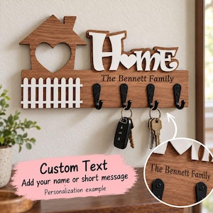 May include: A wooden wall-mounted key holder featuring a house silhouette with a heart cutout, a white picket fence, and the word "Home" in white lettering. It includes four black metal hooks, with keys hanging from two, and the text "The Bennett Family" engraved on the wood.