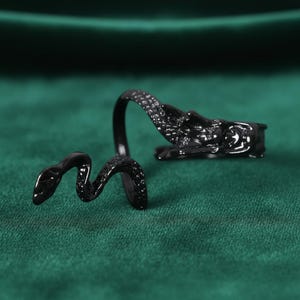 May include: A black snake ring with a detailed design. The ring features a snake wrapping around the finger, with the head and tail meeting. The ring is made of a shiny, dark material and is set against a green background.