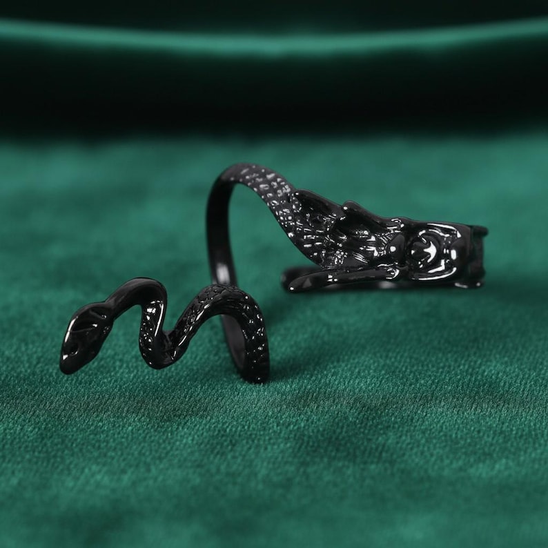 May include: A black snake ring with a detailed design. The ring features a snake wrapping around the finger, with the head and tail meeting. The ring is made of a shiny, dark material and is set against a green background.