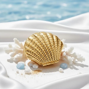 Könnte beinhalten: Ein goldfarbenes, muschelförmiges Kompaktpuder mit strukturierter Oberfläche, das auf weißem Satin liegt. Das Kompaktpuder ist geöffnet und zeigt ein kleines Innenleben. Dekorative Korallen, kleine Perlen sowie blaue und weiße Steine umgeben das Kompaktpuder.