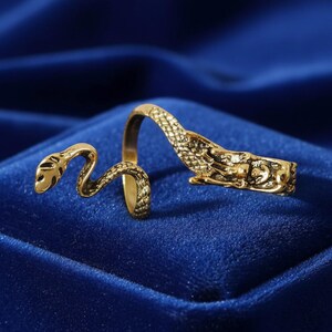 May include: A gold-colored ring in the shape of a snake. The ring wraps around the finger with the snake's head and tail meeting. The ring is set against a blue background.