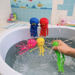 Puede incluir: Una colección de medusas de juguete coloridas en una bañera. Las medusas son azules, rojas, rosas y amarillas, con largos tentáculos. Una mano aprieta un juguete verde, creando un chorro de agua. El fondo presenta una pared de azulejos azules y blancos.