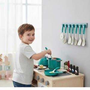 May include: A child plays with a wooden toy kitchen set. The set includes a teal pot, pan, and kettle, along with various cooking utensils hanging on the wall. The child is stirring food in the pot with a spoon.