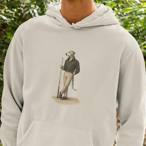 Puede incluir: Una sudadera con capucha color crema con un bolsillo de canguro. La sudadera presenta un gráfico de un perro vestido con una camisa negra y pantalones color caqui, sosteniendo un rifle. La persona de la imagen lleva una gorra verde y pantalones color caqui.